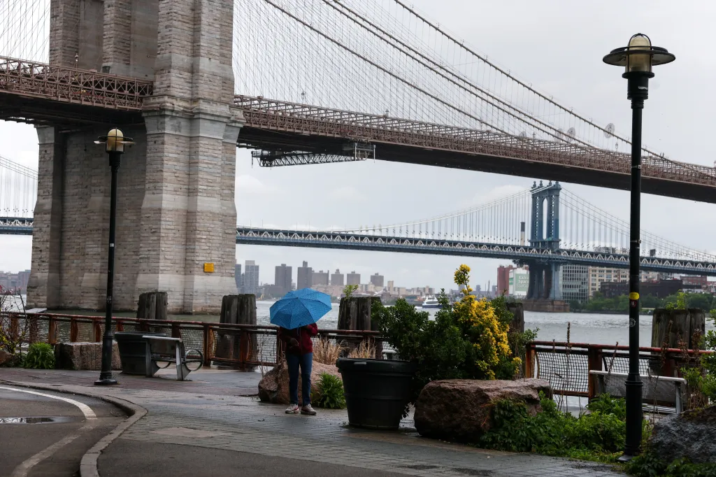 A person with a blue umbrella stands near the Brooklyn Bridge during rainy weather in New York City.