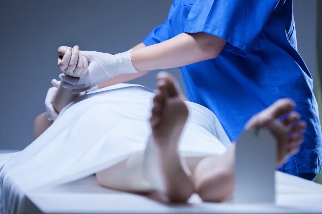 A morgue worker in blue scrubs places an identification tag on the toe of a deceased person on a slab.