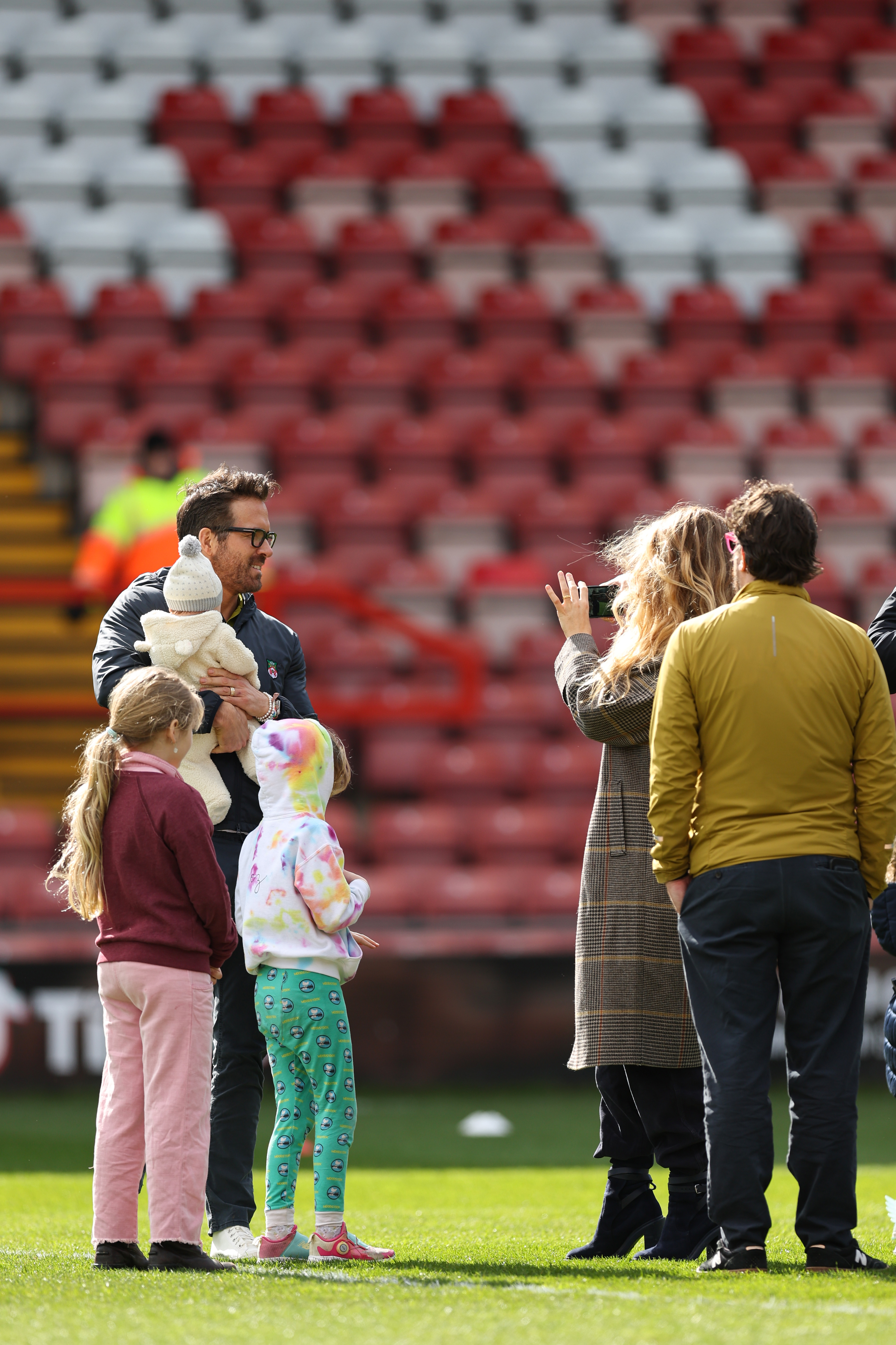 Ryan Reynolds holds his infant, while Blake Lively captures photos, standing on a football pitch with their children and another man.