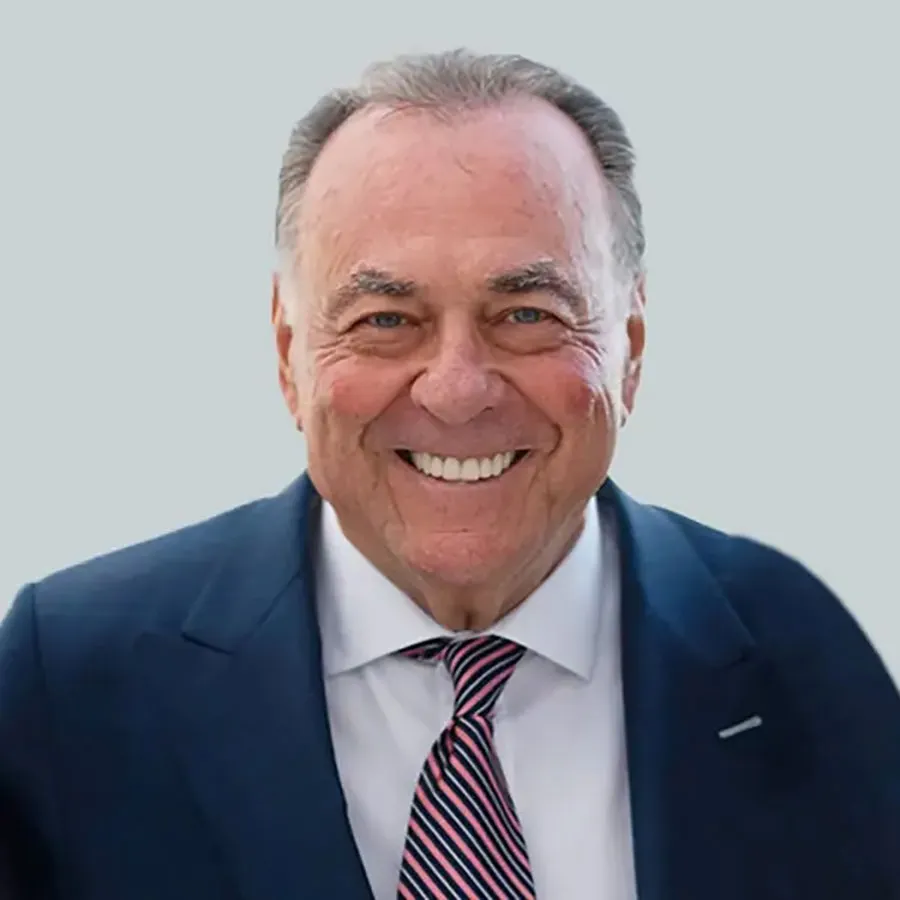 Portrait of Stephen Siegel, a man with gray hair, blue eyes, and a broad smile, wearing a navy suit and a pink and navy striped tie.