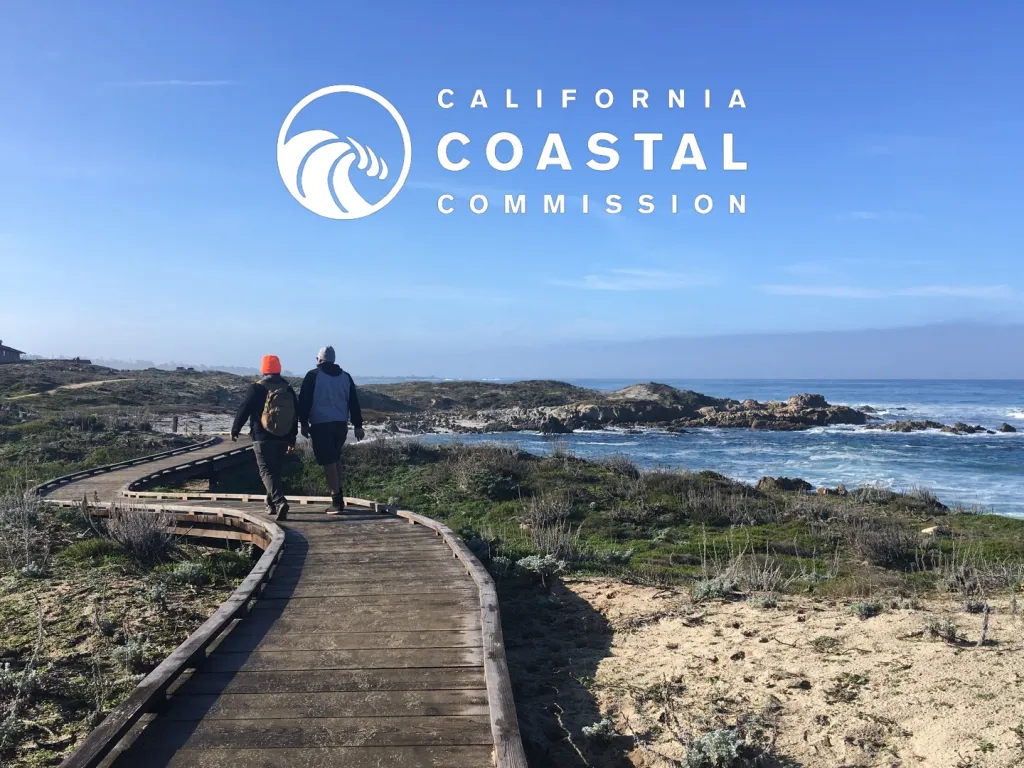 Two people walk on a wooden boardwalk along the California coast under the California Coastal Commission logo.