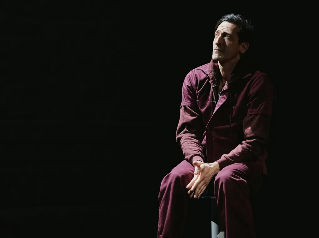 Adrien Brody in a maroon jumpsuit, sitting on a chair, looking up.