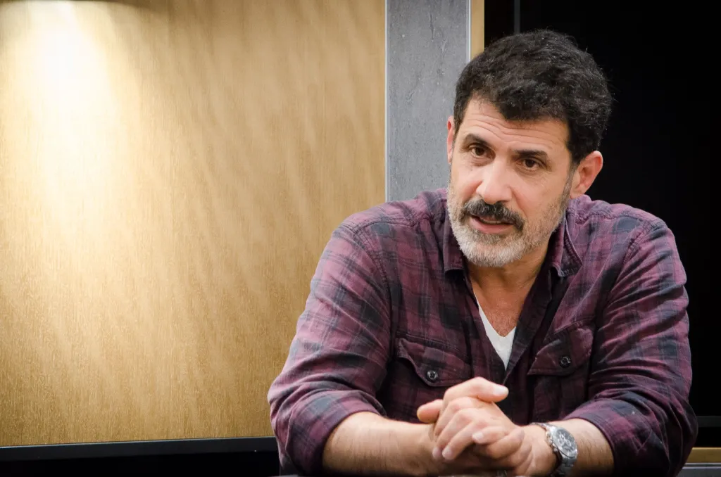 A man with dark hair, a mustache, and a gray beard wearing a plaid shirt and a watch, with his hands clasped, looks to the right of the frame.