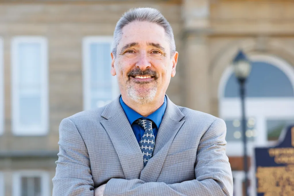 Tracey Powell, a man with gray hair and a beard, smiling and wearing a blue shirt and gray suit with his arms crossed, standing in front of a stone building.