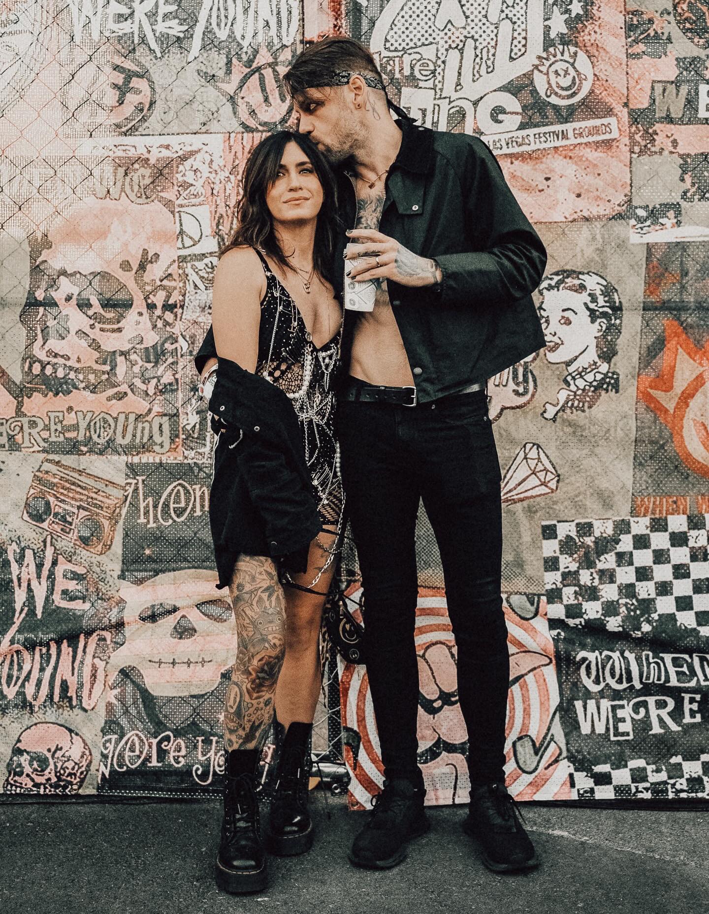 A tattooed man in black kisses a tattooed woman's head, both standing in front of a chain-link fence covered in posters and graffiti.