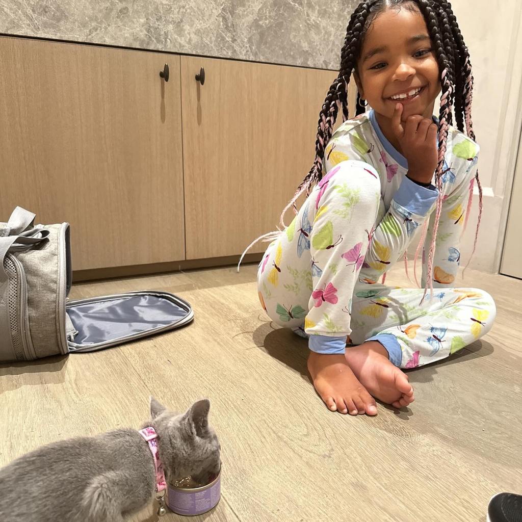 A young girl in butterfly pajamas smiling at a cat eating from a can.
