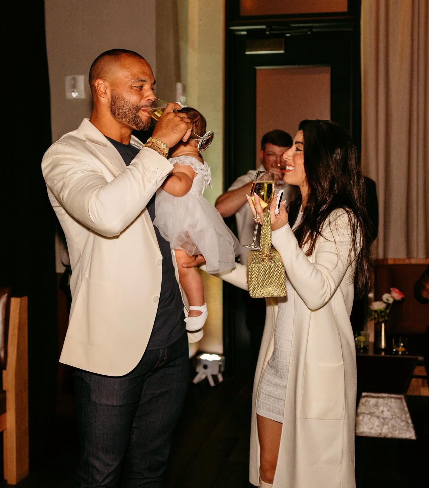 Sarah Jane Ramos and Dak Prescott, holding a baby, raising champagne glasses.