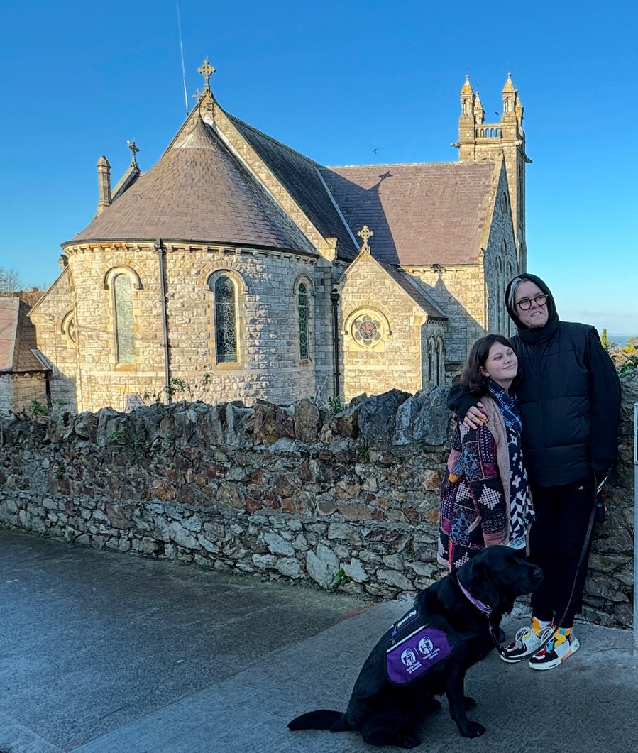 Rosie O'Donnell and her child Clay stand with a black dog in front of a stone church.