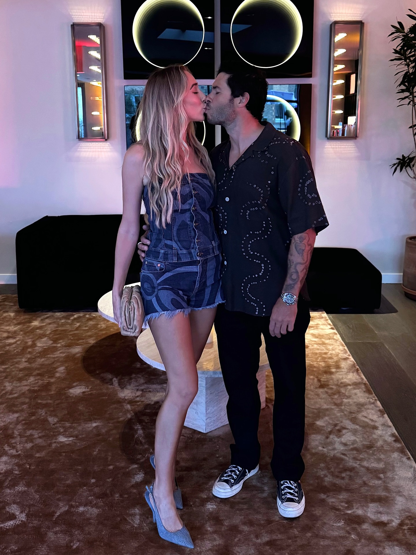 A woman in a denim outfit and a man in a black shirt with white patterns kissing.