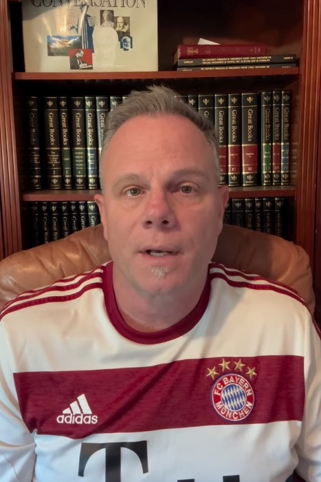 Dr. Joseph Komrosky in a white and red striped t-shirt with a Bayern München logo, sitting in front of a bookshelf.