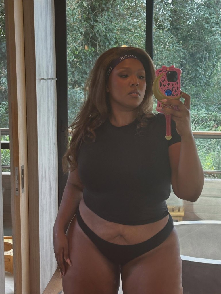 Lizzo posing for a mirror selfie in a black top and underwear while holding a phone with a pink, moon-shaped case.