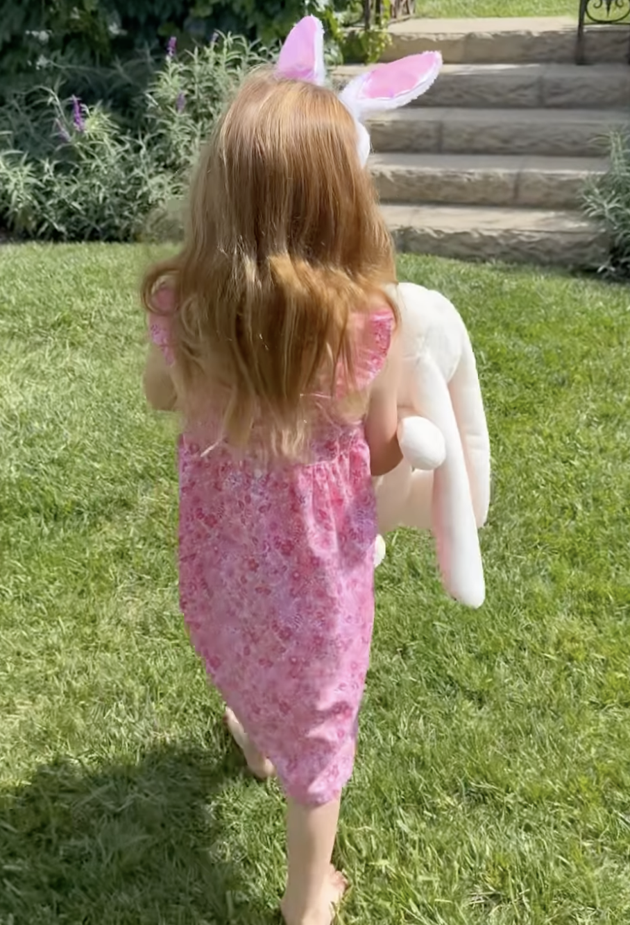 A child with long blonde hair and pink bunny ears from behind, wearing a pink floral dress and carrying a large white plush bunny.