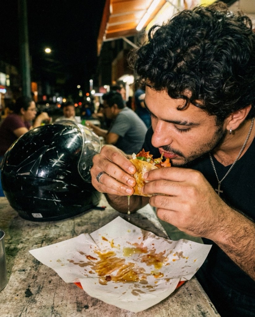 Man with curly hair eating a taco at night.