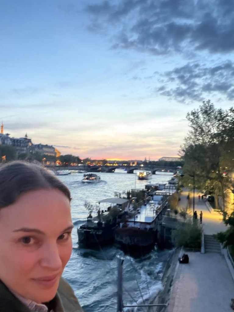 Natalie Portman smiling in a selfie overlooking the Seine River in Paris at dusk.