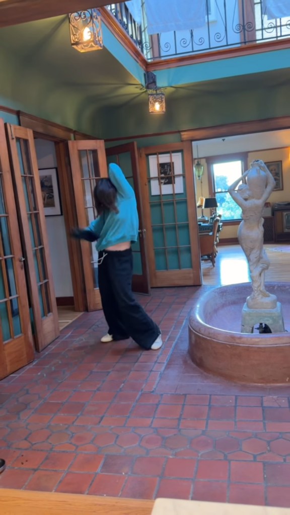 A person in a blue hoodie and black pants dancing in a room with terracotta tile flooring and a statue fountain.
