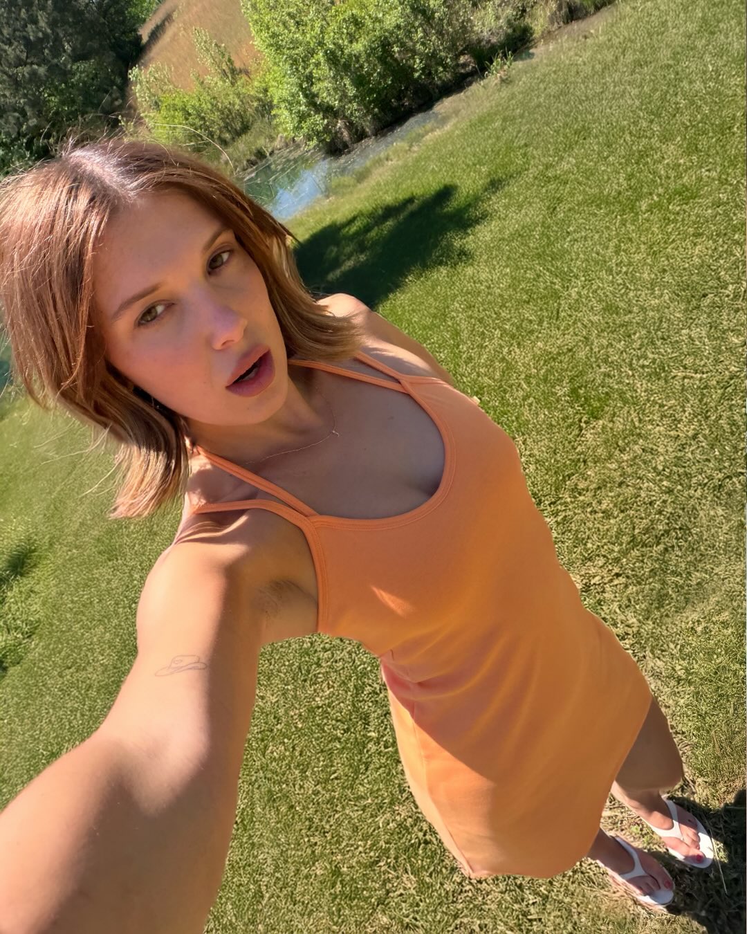 Millie Bobby Brown posing in an orange dress on green grass.