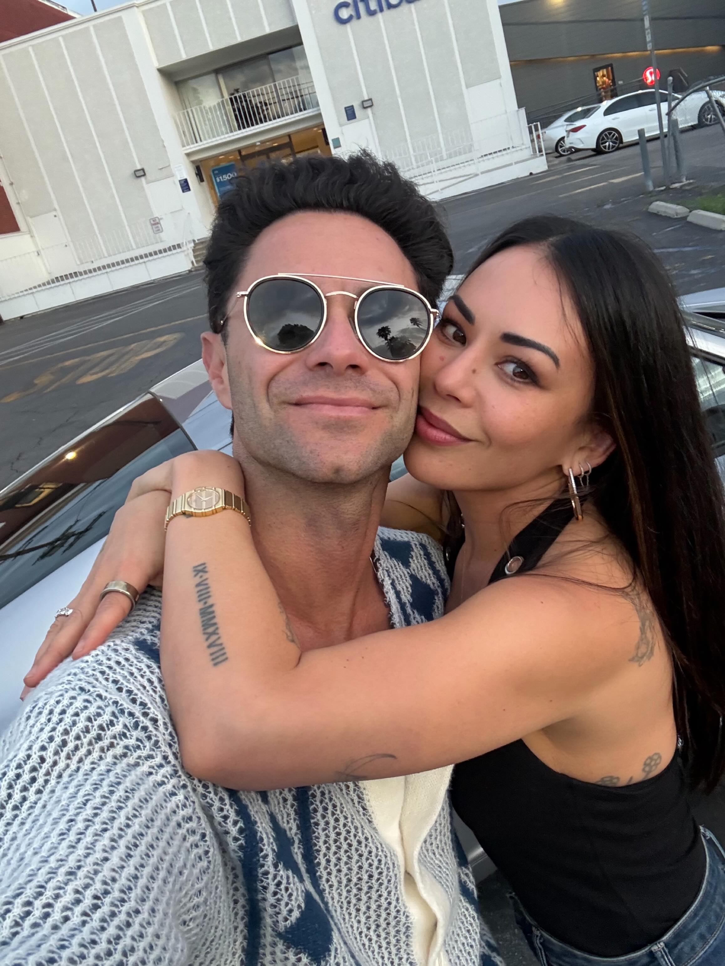 Janel Parrish and Sasha Farber embracing, with Farber wearing sunglasses and Parrish with her head resting on his shoulder.