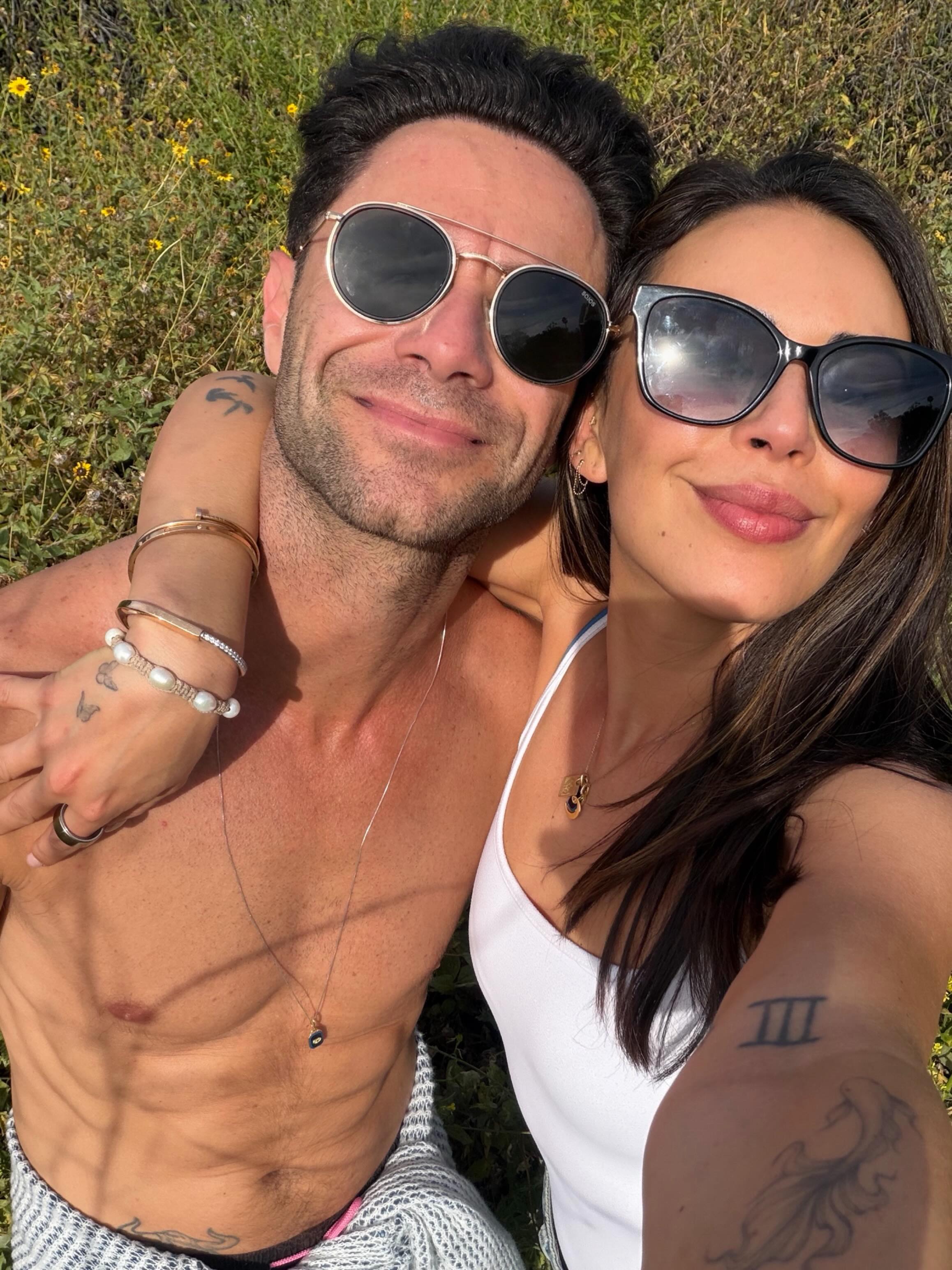 Janel Parrish and Sasha Farber pose for a selfie, smiling in sunglasses while surrounded by green foliage with yellow flowers.