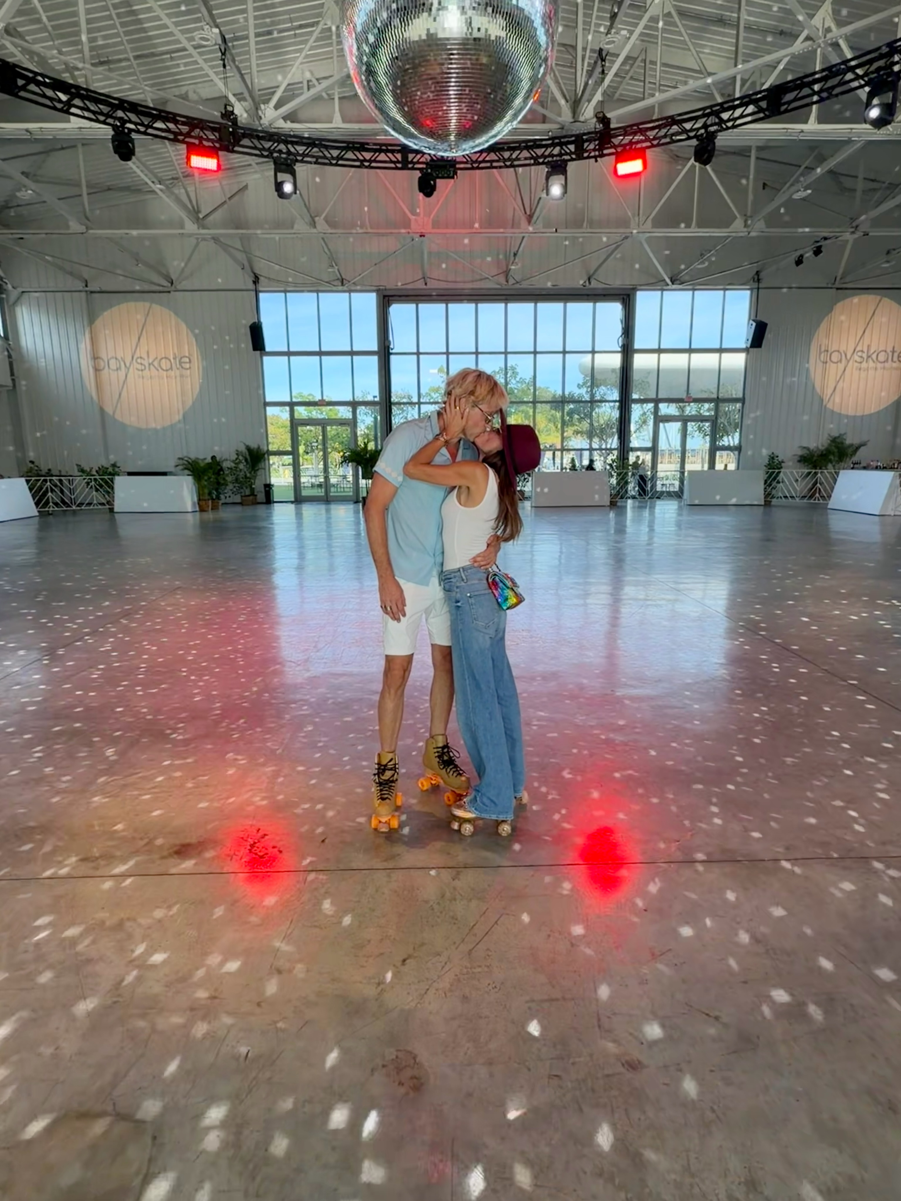 A man and woman wearing roller skates kiss in a roller rink with a disco ball above them.