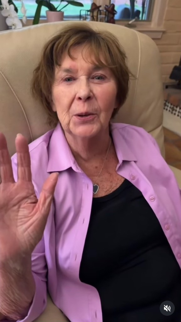 A woman in a lilac shirt and black top, with a silver heart pendant, is seated in a beige chair, gesturing with her left hand.