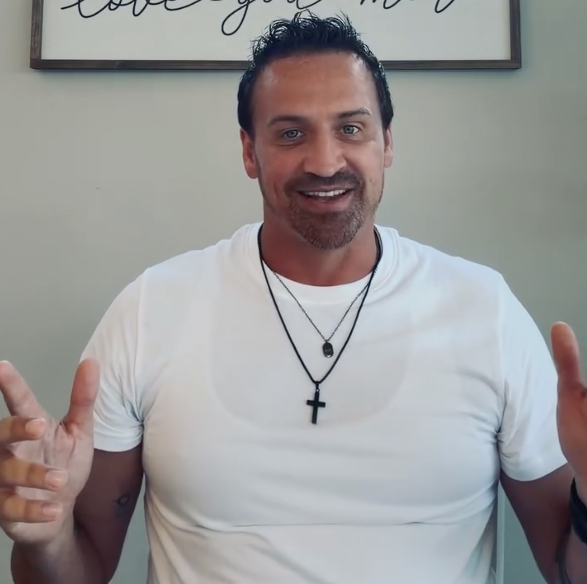 Ryan Lochte with a beard, wearing a white t-shirt and two black necklaces, smiling and talking.