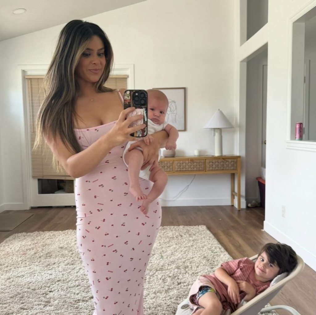 A woman in a pink dress holding a baby, taking a mirror selfie while a young boy reclines in the foreground.