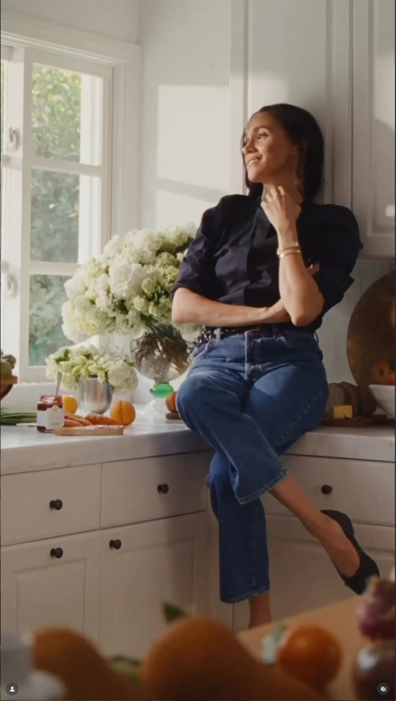 Meghan Markle, I a black top and blue jeans, sitting on a kitchen counter, surrounded by fruits, vegetables, flowers, and jars of preserves.