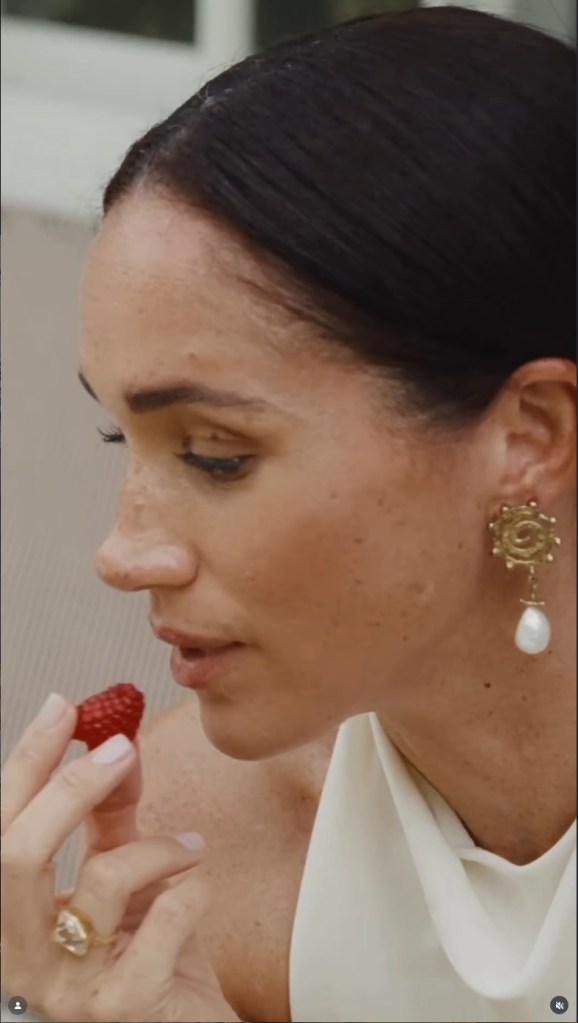 Meghan, Duchess of Sussex,wearing a white dress, holds a raspberry to her mouth.