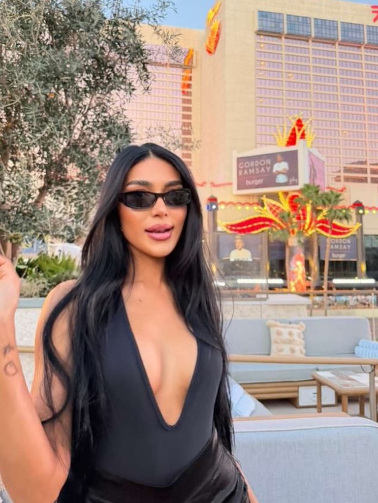 Thais Ramone posing in a black top and sunglasses in front of a building with a Gordon Ramsay Burger sign.
