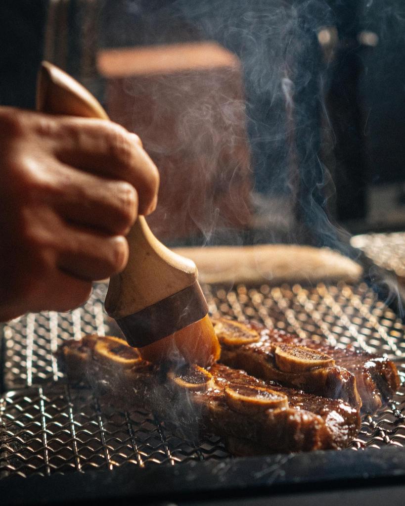 A hand brushes sauce onto short ribs cooking on a grill, surrounded by smoke.