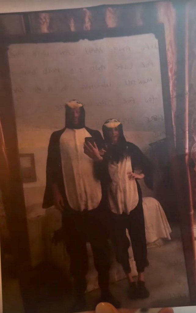 Meghan Markle and Prince Harry wearing penguin onesies, reflected in a mirror with writing on it.