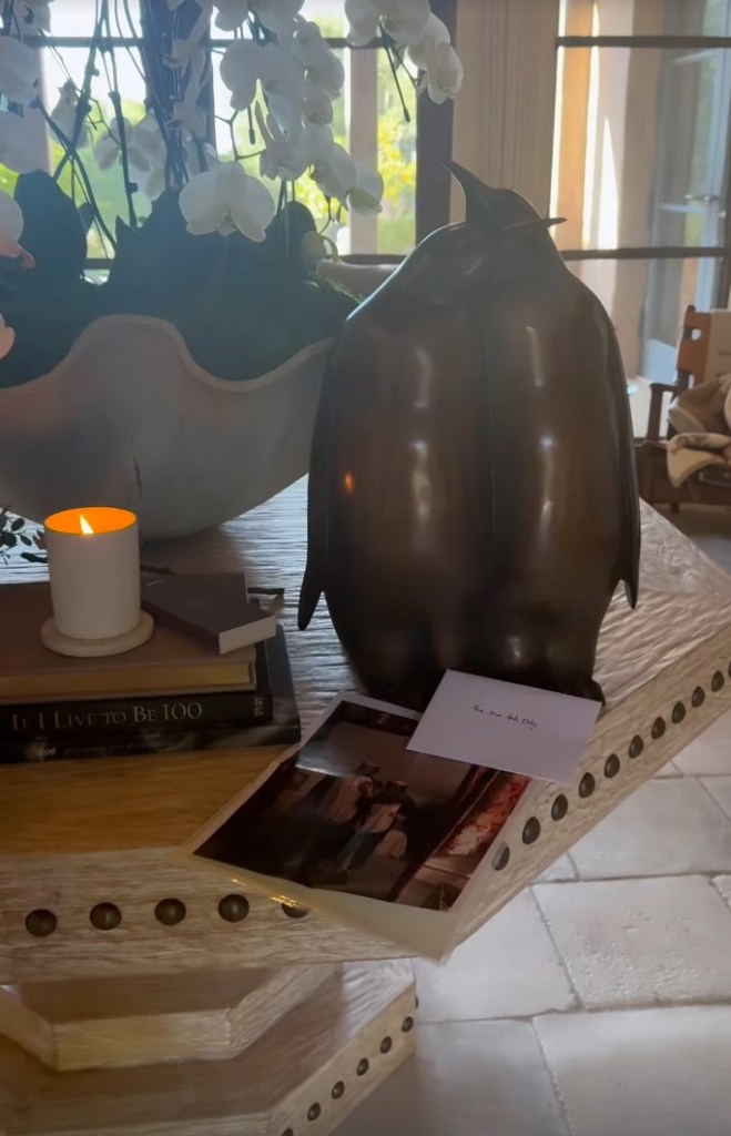 A bronze penguin sculpture next to a lit candle, a book, a photograph, and a card that reads