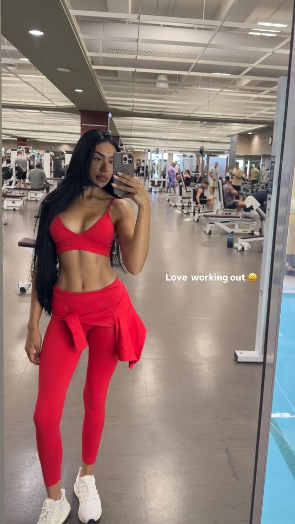 Thais Ramone posing for a mirror selfie in a gym, wearing a red sports bra and leggings.