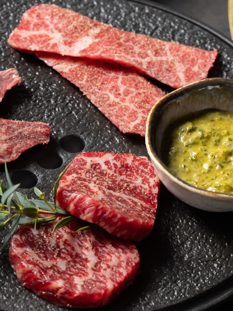 Several cuts of raw, marbled beef seasoned with pepper, with a side of green sauce and a sprig of greenery, on a black textured surface.