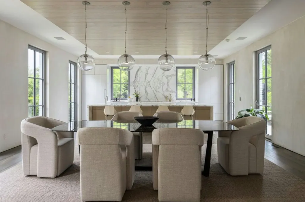 A dining room with a glass table and four cushioned chairs in the foreground, and a kitchen with white marble countertops and white cabinets in the background.