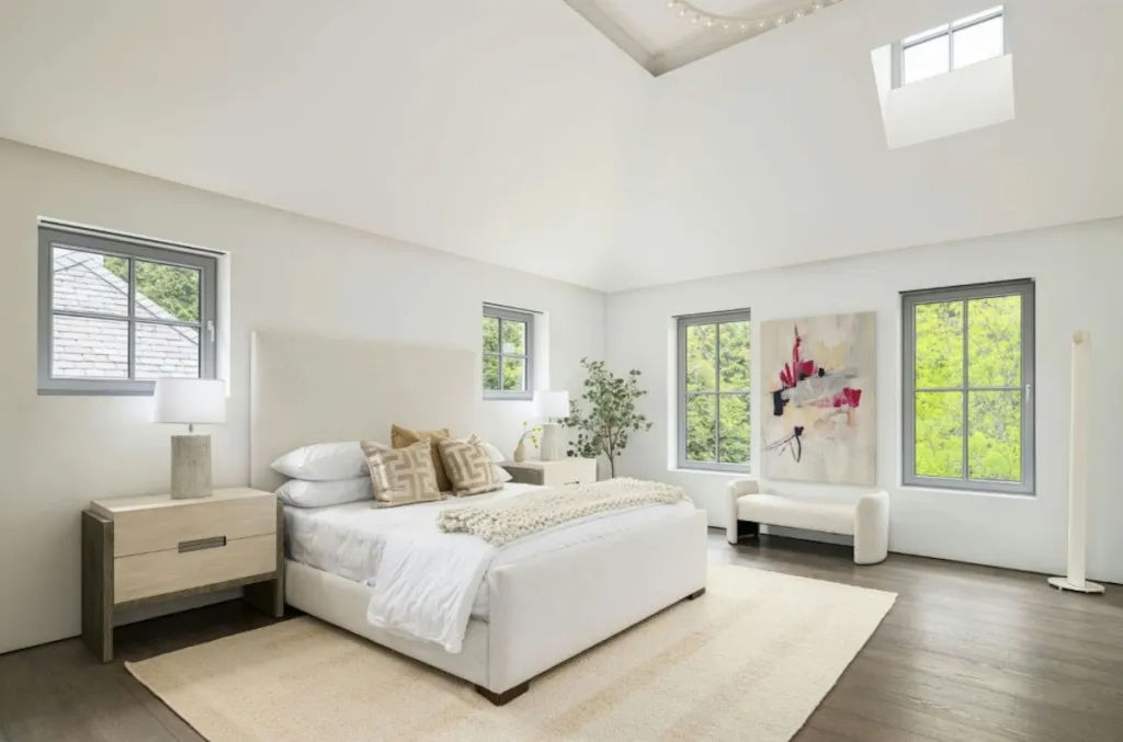 Modern bedroom with a white upholstered bed, light wood nightstands, and large windows with views of greenery.