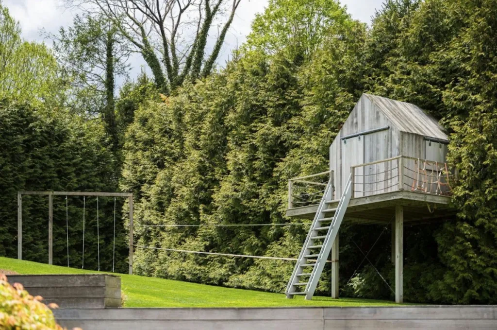 A rustic wooden treehouse with a ladder, balcony, and rope netting stands amidst a lush green lawn and dense trees, with a swing set visible in the background.