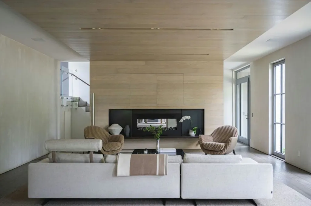 A modern living room with a large light wood fireplace and two beige armchairs facing a white sectional sofa.