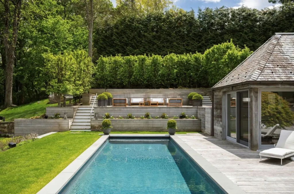 A narrow, rectangular swimming pool bordered by a wooden deck and grass, with tiered landscaping leading up to a lush green hillside.