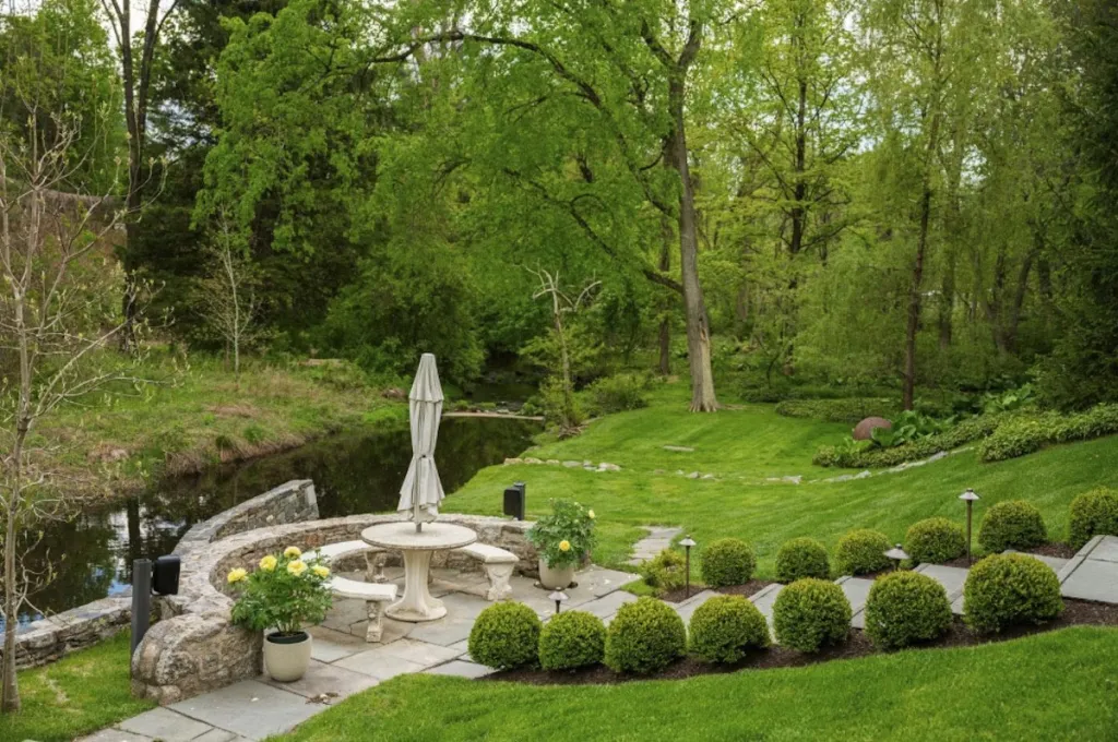 Patio with a table and umbrella next to a body of water, surrounded by green trees and trimmed bushes.