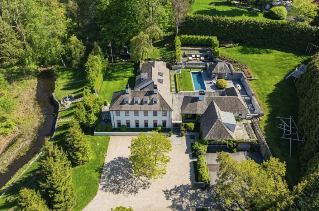 Aerial view of a large white house with a gravel driveway, surrounded by green lawns, trees, and a swimming pool.