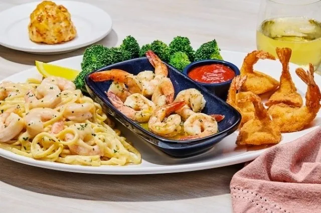 A plate of shrimp scampi pasta, garlic shrimp in a black dish, and breaded shrimp with broccoli, cocktail sauce, and a lemon wedge, with a Cheddar Bay Biscuit and a glass of white wine in the background.