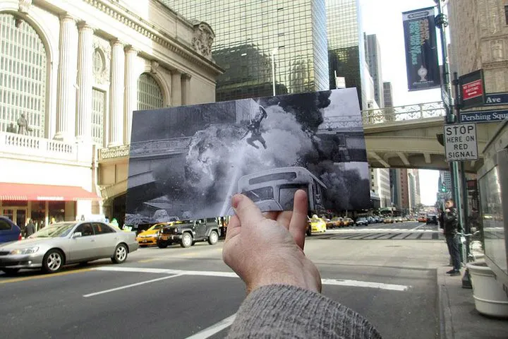 A hand holds a black and white movie still of an explosion with a superhero mid-air in front of a modern city street.