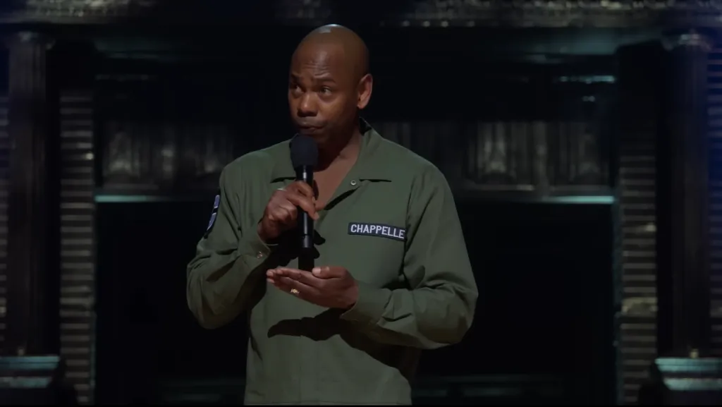 Dave Chappelle performing stand-up comedy on stage, holding a microphone.