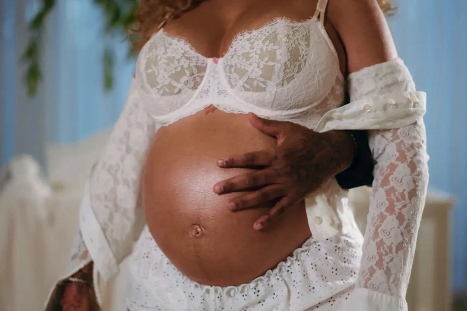 A pregnant person in a white lace bra and shorts, with a partner's arm around their waist, cradling their belly.