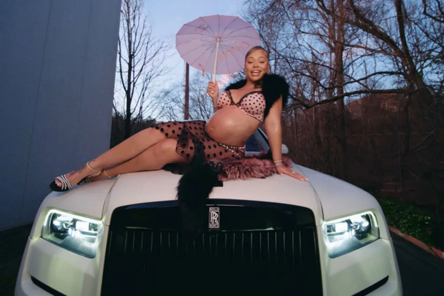 Latto, pregnant and wearing a polka-dot bra and skirt with a fur boa, lies on the hood of a white Rolls Royce while holding an umbrella.