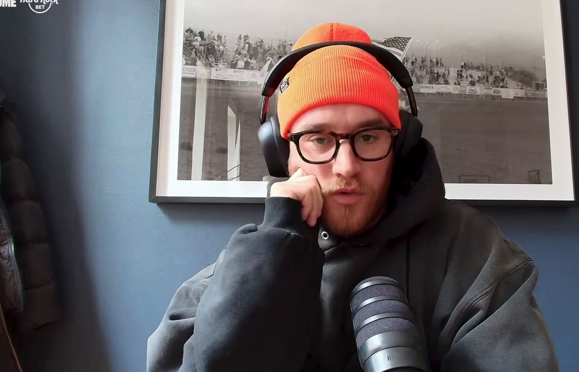 A man with an orange beanie, headphones, and glasses speaking into a microphone.