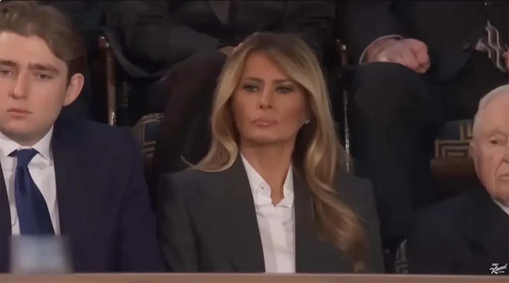 Melania Trump looking ahead, seated between a young man and an older man.