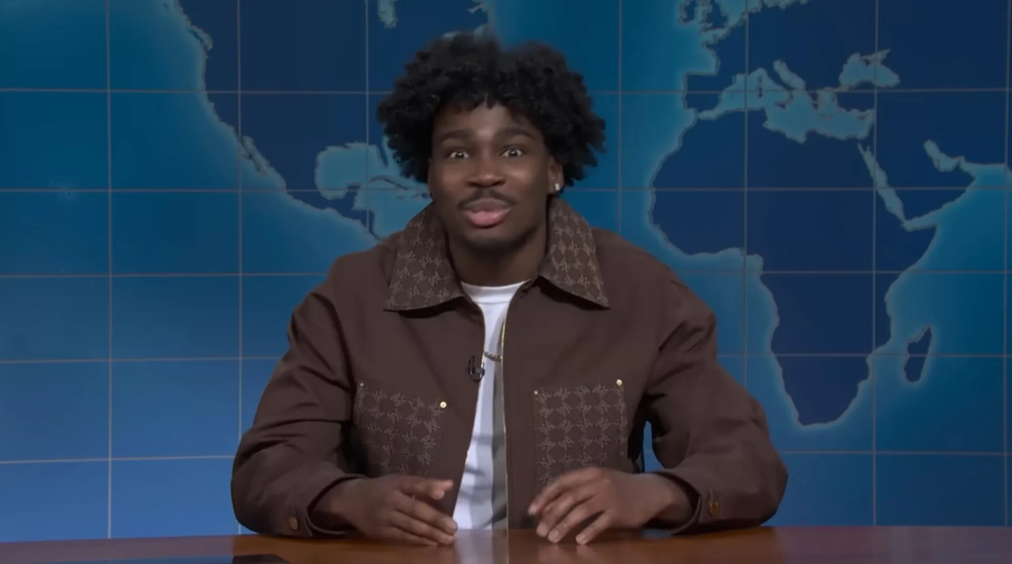 Comedian Kam Patterson standing in front of a blue world map, wearing a brown jacket with patterned lapels, speaking into a microphone.