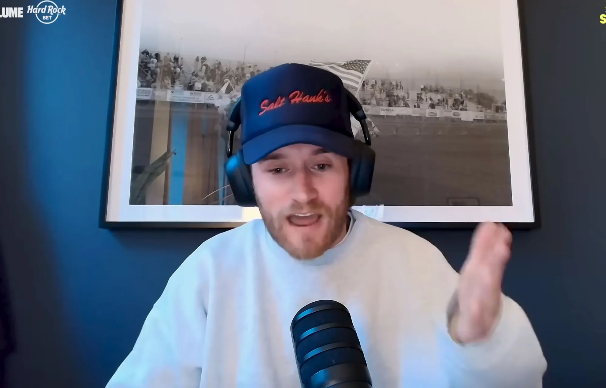 West Wilson in a blue baseball cap, headphones, and a white sweatshirt, speaking into a microphone.